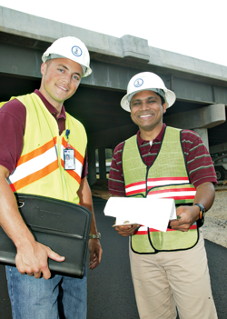 Two engineers, wearing hardhats, review documents at a job site.